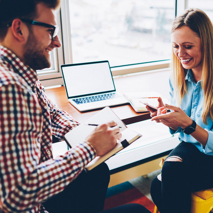 Besprechungssituation Ein Mann mit Notizblock und Stift und eine Frau mit Handy sitzen sich gegenüber und lächeln entspannt. An ihrer Seite steht ein Tisch mit geöffnetem Laptop. Die Stühle sind gelb.